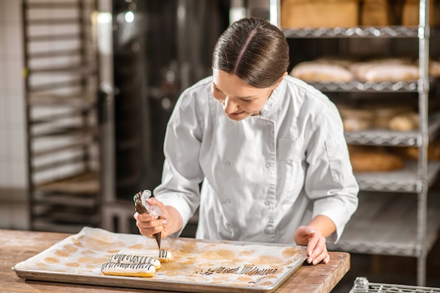 Trabajo favorito. Interesada mujer atenta pastelero en chaqueta blanca decorar canutillos con patrón de chocolate en la panadería