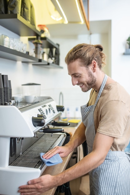 Trabajo favorito. Sonriente joven en delantal con servilleta azul limpiando la superficie de la máquina de café de buen humor