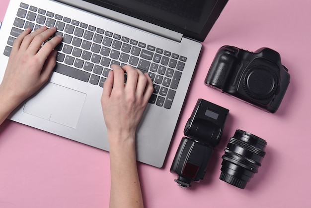 El trabajo del fotógrafo, retoque fotográfico. Equipo fotográfico, manos femeninas escribiendo en el teclado del portátil sobre fondo rosa pastel, vista superior, concepto de trabajo independiente, plano