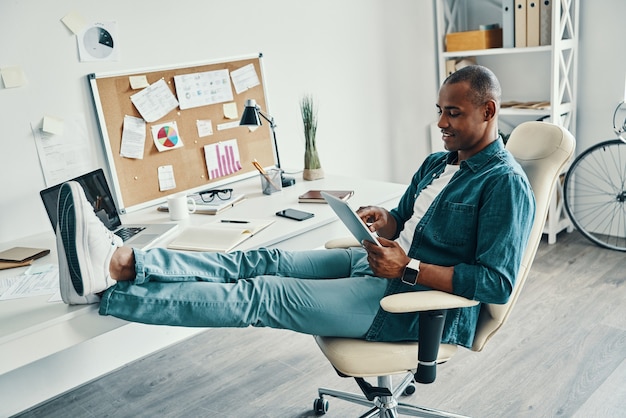 Trabajo independiente. Apuesto joven africano en camisa con tableta digital y sonriendo mientras está sentado en la oficina