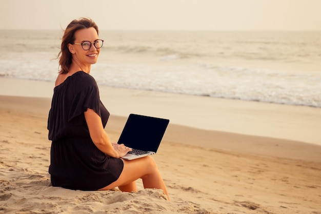 Trabajo independiente y remoto. mujer de negocios en un elegante traje de verano de negocios con una computadora portátil sentada en la arena de la palma de la costa del océano tropical. estudiante con anteojos educación en línea en el extranjero