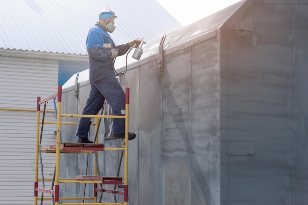 Trabajo industrial Cebado de productos metálicos de la pistola del compresor Un trabajador con un mono y un respirador pinta el cuerpo de un remolque de camión o un automóvil de metal