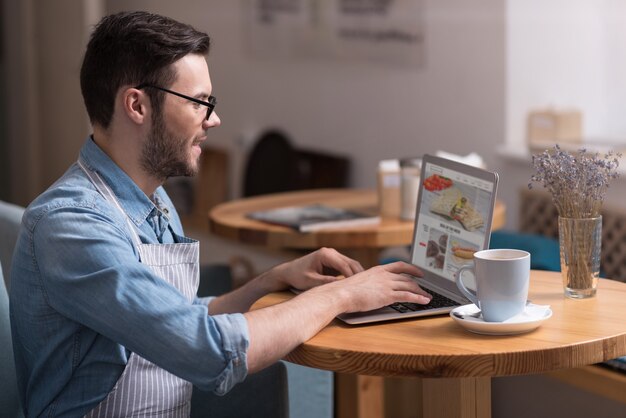 Trabajo progresivo. Feliz apuesto joven sonriendo y usando la computadora portátil mientras está sentado en la mesa.