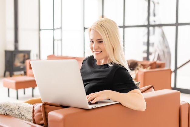 Trabajo remoto en casa, autónomo durante la cuarentena de covid-19. Mujer sonriente sentada y trabajando en una laptop en el acogedor salón interior, espacio libre.