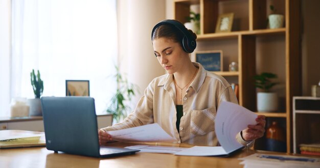 Trabajo remoto desde casa documentos y mujer con auriculares escuchando música y portátil Persona independiente y emprendedora con auricular de PC y lectura en línea para papeleo de proyecto o conexión
