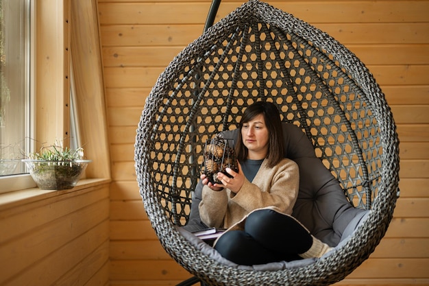 Trabajo remoto y escapar al concepto de la naturaleza La mujer se sienta en un columpio de silla de huevo y sostiene un jarrón de conos en una pequeña cabaña de madera