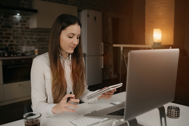 Trabajo remoto. Una mujer caucásica con auriculares trabajando remotamente en su computadora portátil. Una chica con una camisa blanca en busca de sus notas durante una sesión informativa de negocios en línea en su acogedor lugar de trabajo en casa.