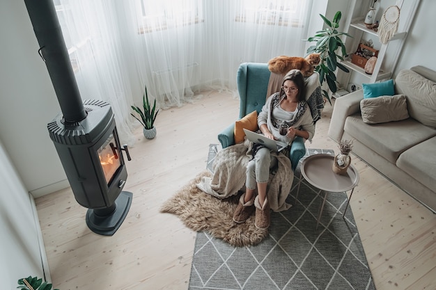 Trabajo remoto. Mujer joven con ordenador portátil en un acogedor sillón y cuadros sentado junto a la chimenea con un gato doméstico, bebiendo té en casa