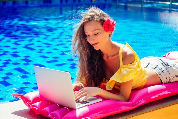 Trabajo remoto trabajo de ensueño mujer joven sentada en un colchón rosa inflable en la piscina tomando el sol y trabajando en una computadora portátil chica comiendo frutas exóticas mango tropical piña y fruta del dragón