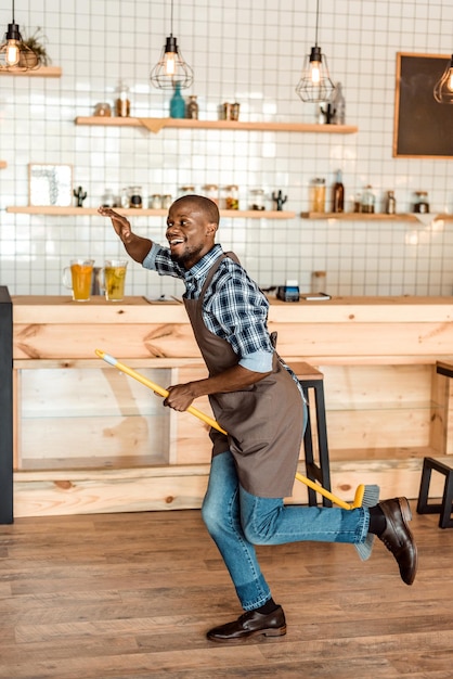 Trabalhador afro-americano bonito se divertindo e pulando com a vassoura no café