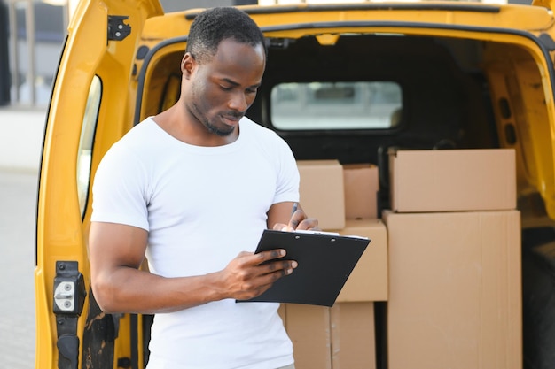 Trabalhador afro-americano descarregando caixas de van ao ar livreConceito de serviço de mudançaFuncionário masculino da empresa de mudanças faz inventário de caixas embaladas e grava dados na área de transferência