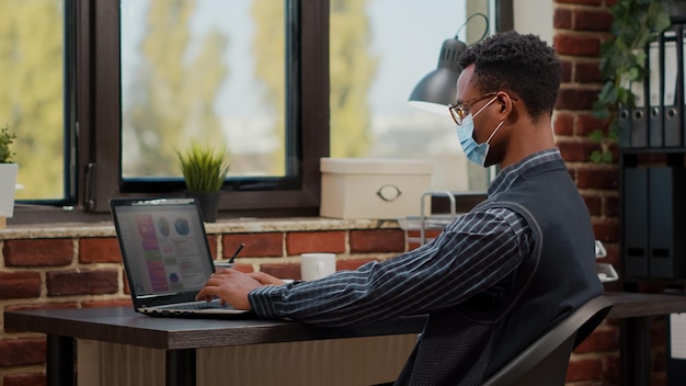 Trabalhador americano africano analisando o crescimento comercial no laptop no escritório de inicialização. Funcionário da empresa usando máscara facial e trabalhando no desenvolvimento de marketing com relatório de vendas no computador.