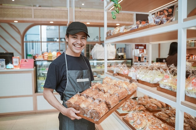 Trabalhador asiático sorridente e bonito na padaria segurando uma bandeja de pão