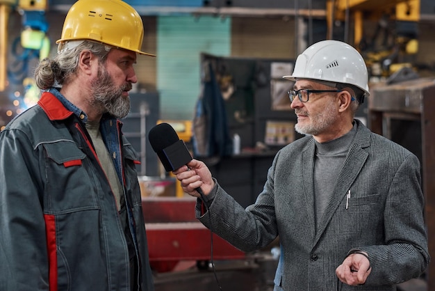 Trabalhador barbudo maduro de uniforme conversando com jornalista de TV durante entrevista na fábrica