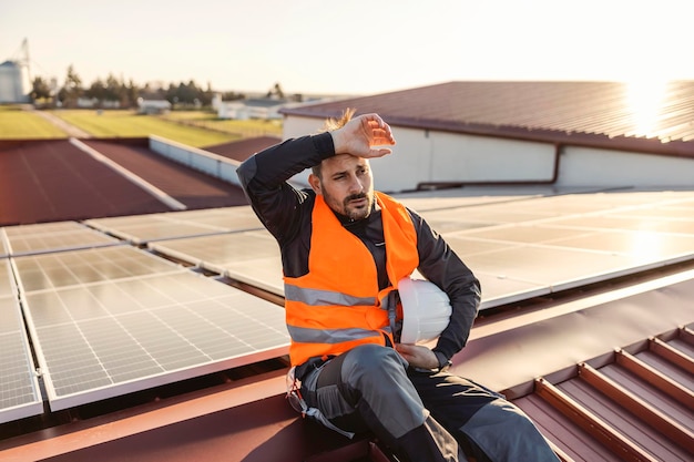 Trabalhador cansado está sentado no telhado com painéis solares instalados e suor arrebatador
