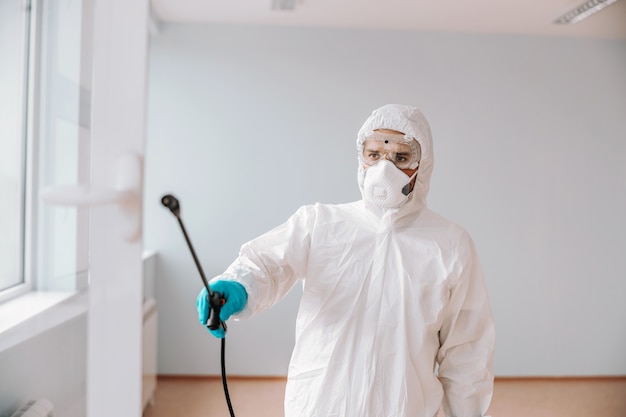 Trabalhador com uniforme esterilizado, com máscara e luvas borrifando desinfetante com janela na escola.