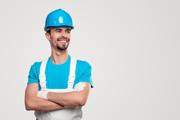 Trabalhador da construção civil adulto positivo, barbudo, com capacete azul e macacão branco, e luvas em pé com os braços cruzados e olhando para longe com confiança contra um fundo branco