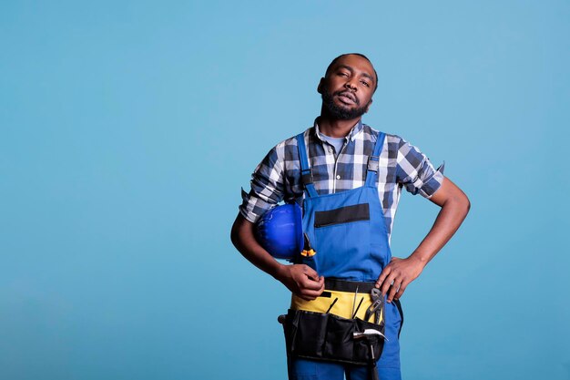 Trabalhador da construção civil americano africano olhando seriamente para a câmera no estúdio atirou contra um fundo azul. Trabalhador da construção civil no final do dia de trabalho segurando o capacete debaixo do braço.