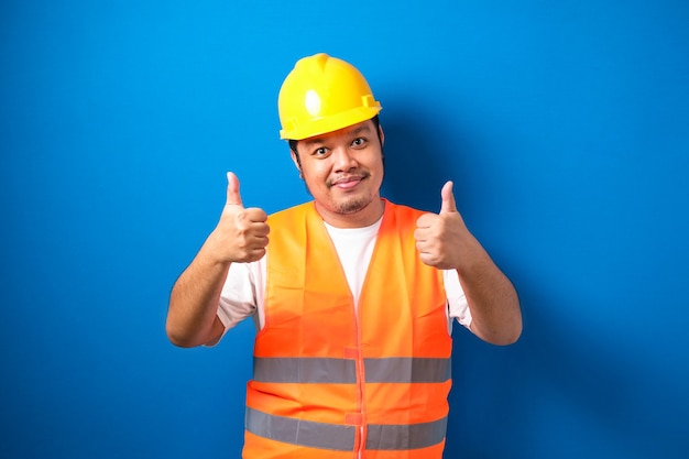 Trabalhador da construção civil asiático gordo usando colete de segurança laranja e capacete mostrando o polegar para cima a placa sorrindo