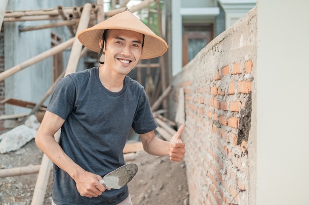 Trabalhador da construção civil asiático sorrindo para a câmera usando um boné com o polegar para cima enquanto segura a colher