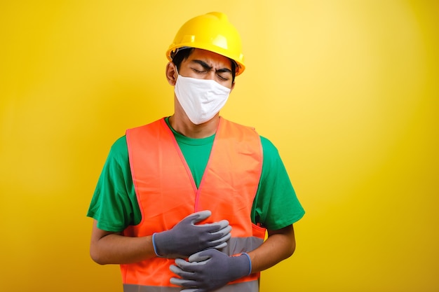 Trabalhador da construção civil asiático usando capacete e máscara protetora contra um fundo amarelo com a mão na barriga por causa de indigestão, doença dolorosa, mal-estar. conceito de dor.