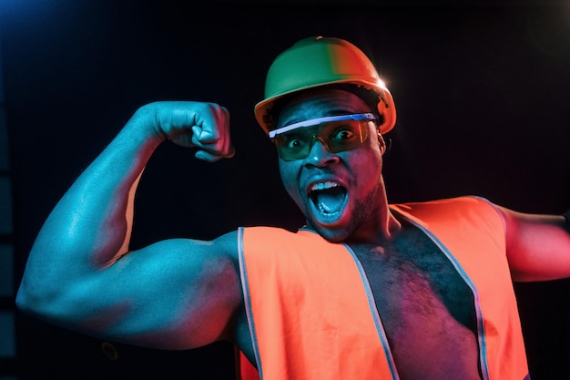 Trabalhador da construção civil de uniforme e capacete de segurança iluminação neon futurista jovem afro-americano no estúdio