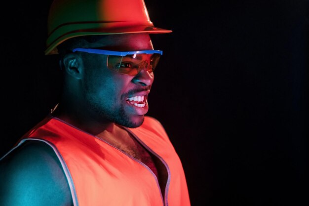 Trabalhador da construção civil de uniforme e capacete de segurança Iluminação neon futurista Jovem afro-americano no estúdio