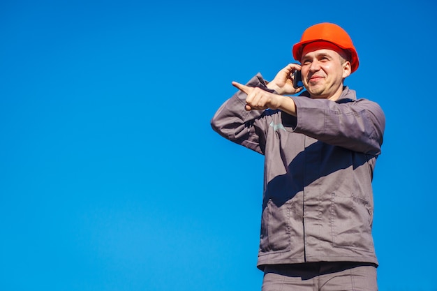 Trabalhador da construção civil em um capacete contra o céu azul, falando ao telefone.