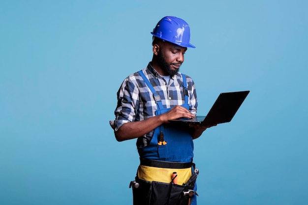 Trabalhador da construção civil em uniforme de trabalho e capacete usando computador portátil moderno. Supervisor de canteiro de obras masculino profissional usando um cinto de ferramentas ao usar um computador em um fundo azul.