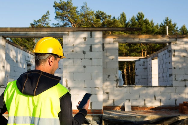 Trabalhador da construção civil fala em um smartphone em um capacete amarelo e colete reflexivo no contexto da construção da casa - as paredes e as aberturas das janelas são feitas de blocos de concreto porosos.