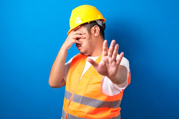 trabalhador da construção civil homem vestindo uniforme e capacete cobrindo os olhos com as mãos e fazendo gesto de pare