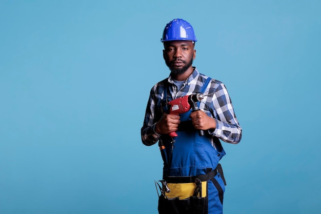 Trabalhador da construção civil profissional com macacão e ferramentas de trabalho contra um fundo azul. Tiro de estúdio homem afro-americano olhando para a câmera segurando a furadeira elétrica sem fio.