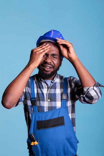 Trabalhador da construção civil sofrendo de uma forte dor de cabeça ao remover o capacete de proteção. Empreiteiro estressado pelo excesso de trabalho tocando sua testa com expressão de dor contra o fundo azul.