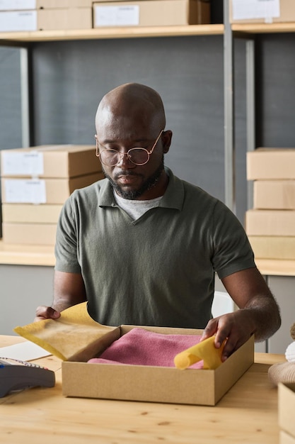 Trabalhador de armazém africano embalando pedido de cliente em caixa de papelão enquanto está sentado à mesa no storag