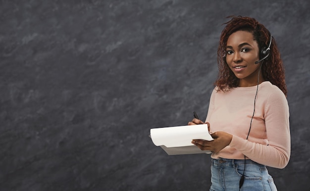 Trabalhador de call center muito afro-americano com fones de ouvido. Mulher fazendo anotações em fundo cinza