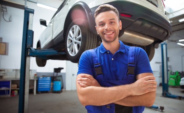 Trabalhador de conserto de automóveis jovem e experiente, bonito e confiante em macacão posando no contexto de carros levantados em uma oficina de automóveis