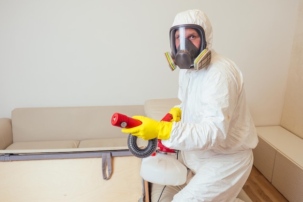 Trabalhador de controle de pragas em uniforme pulverizando pesticidas sob o sofá na sala de estar