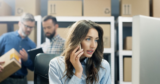 Trabalhador de correios de mulher jovem e bonita falando no celular enquanto trabalha no computador.