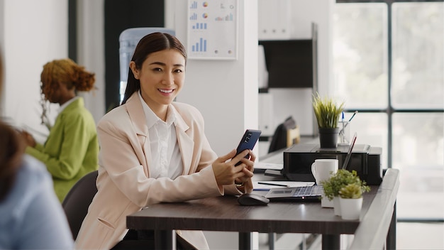 Trabalhador de escritório asiático usando um aplicativo de smartphone no local de trabalho, planejando um relatório on-line antes de fazer uma pausa na mesa. Jovem funcionário navegando na internet e no aplicativo de mídia social no espaço de coworking.