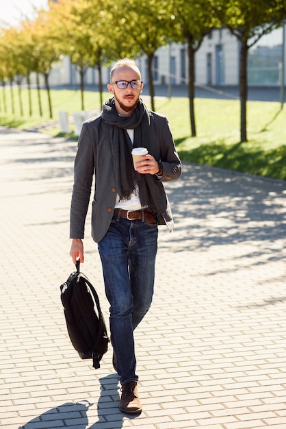 Trabalhador de escritório bonito de terno com lenço segurando uma sacola e bebe café da manhã antes do início do trabalho perto do prédio de escritórios. Jovem empresário, indo para a reunião de negócios.