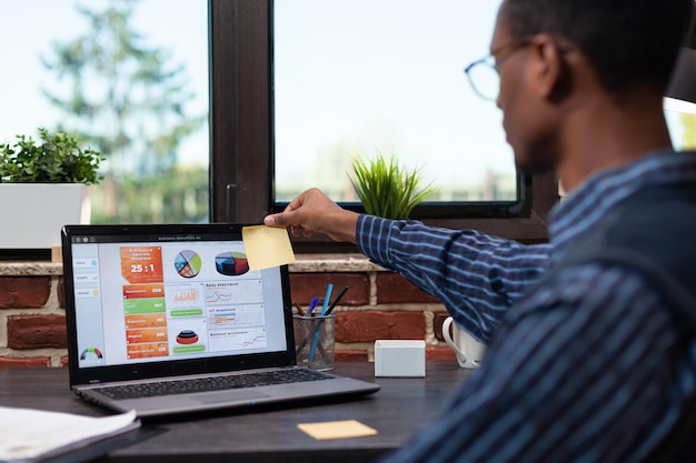 Trabalhador de escritório com óculos deixando adesivo de mensagem de lembrete no canto da tela do laptop com dados de vendas de negócios. empregado de inicialização sentado na mesa colando amarelo post-it na tela do computador portátil.