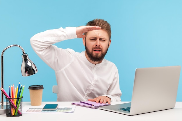 Trabalhador de escritório de homem barbudo ambicioso sentado no local de trabalho com laptop e olhando para longe com a mão acima dos olhos, pesquisando com promoção, explorando o futuro. Tiro de estúdio interior isolado em fundo azul