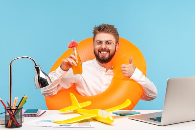 Foto trabalhador de escritório de homem extremamente feliz sentado no local de trabalho com boia salva-vidas de borracha e avião de brinquedo segurando a garrafa de limonada e mostrando os polegares para cima relaxante tiro de estúdio interior isolado em fundo azul