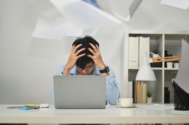 Trabalhador de escritório estressado se sentindo angustiado e ansioso com o prazo de trabalho sentado à mesa com a papelada caindo