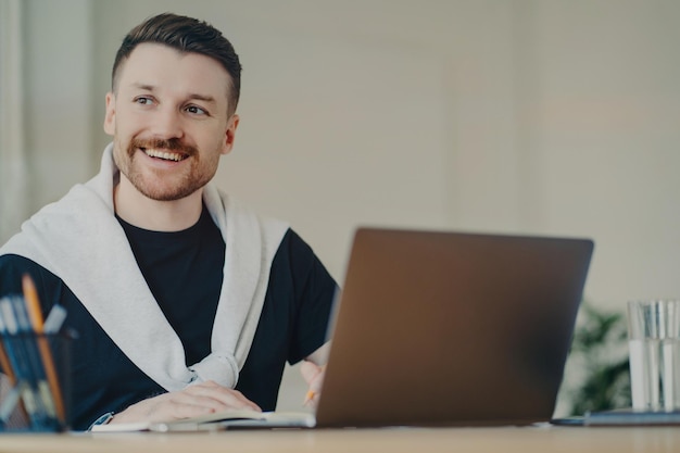 Trabalhador de escritório feliz homem barbudo escreve plano de organização funciona remotamente faz anotações de informações senta na frente do laptop aberto desvia o olhar vestido com uma camiseta casual e um macacão amarrado nos ombros