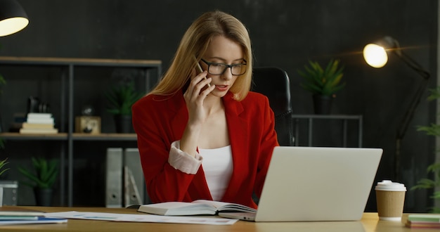 Trabalhador de escritório feminino caucasiano sentado à mesa, falando no telefone celular enquanto trabalhava no computador portátil.