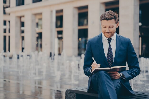 Trabalhador de escritório formalmente vestido, organizando seu dia no caderno, escrevendo tudo enquanto está sentado no banco, fontes trabalhando atrás dele, empresário bonito aproveitando o trabalho ao ar livre no fim de semana