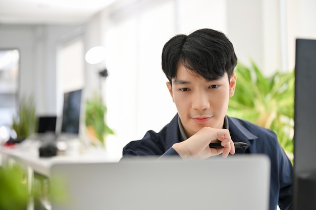 Trabalhador de escritório masculino asiático inteligente com a mão no queixo pensando e olhando para a tela do computador