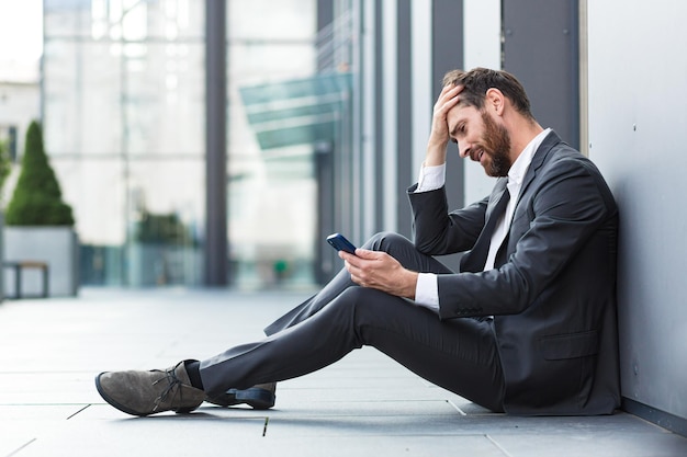 Trabalhador de escritório sentado perto do escritório, ao ar livre, recebendo más notícias, deprimido e cansado, lendo más notícias no telefone online