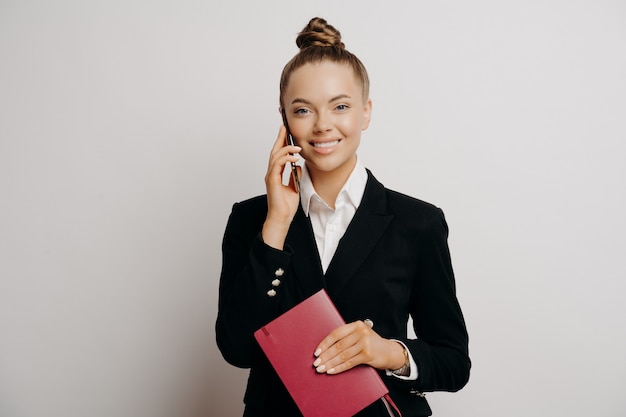 Trabalhador de escritório sorridente, mulher em traje formal preto conversando ao telefone, segurando um caderno vermelho com dados, ouvindo boas notícias enquanto está isolado em um fundo cinza